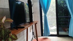 A room with a television on a wooden stand, a sliding glass door with blue curtains, and a concrete floor. - M&K House - Family JUD Room (Tarapoto)