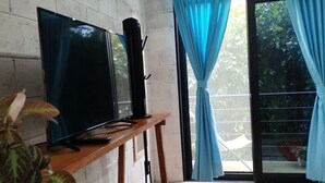 A room with a TV on a wooden stand, a potted plant, a sliding glass door with blue curtains, and a concrete floor. - M&K House - Family JUD Room (Tarapoto)