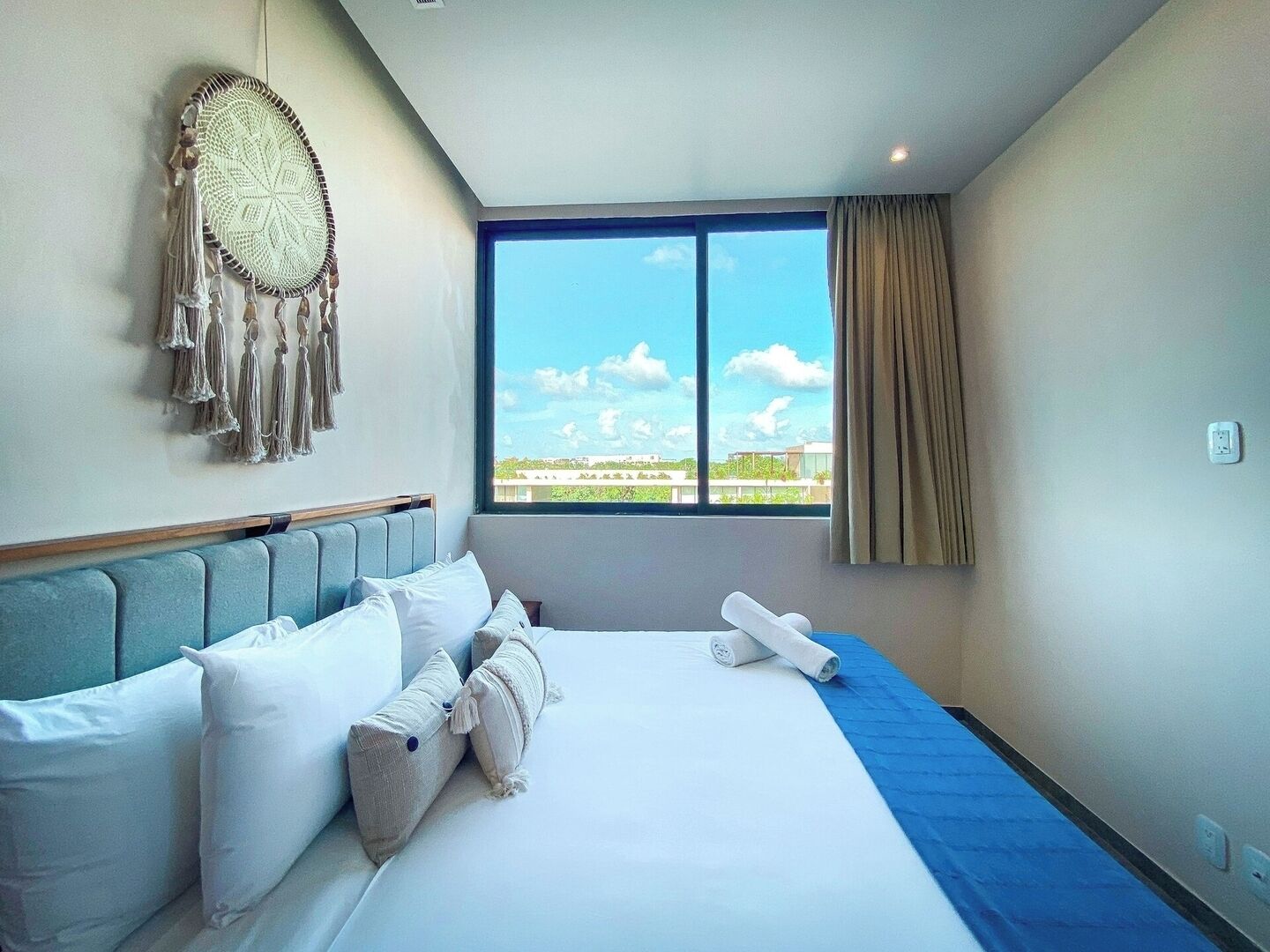 A hotel room with a large bed, a headboard, a dreamcatcher, a view of buildings through the window, and a power outlet on the wall.