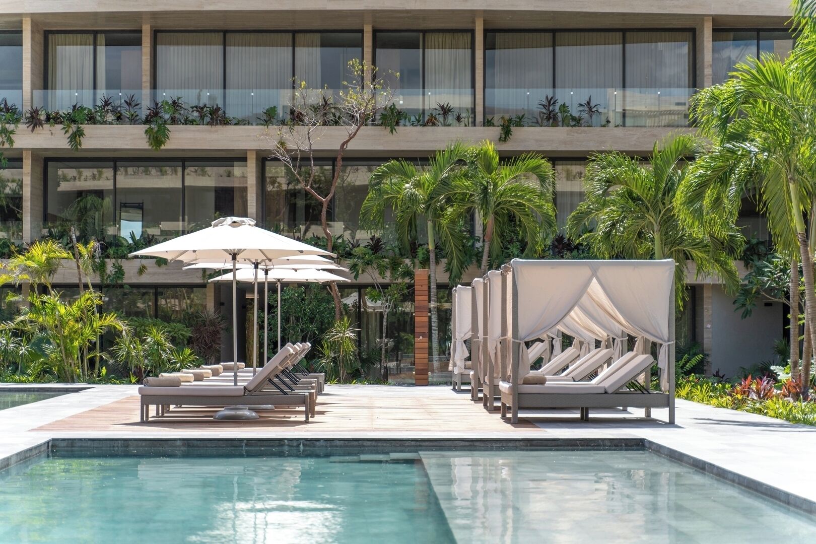 Area piscina con sedie a sdraio e ombrelloni, immersa nel verde e con affaccio su un edificio moderno dotato di balconi.