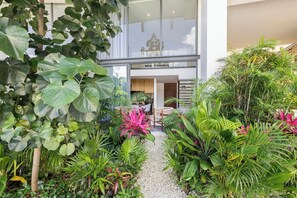 Un bâtiment moderne doté d’une façade en verre, d’un petit espace extérieur aménagé en coin salon et d’une végétation tropicale luxuriante.