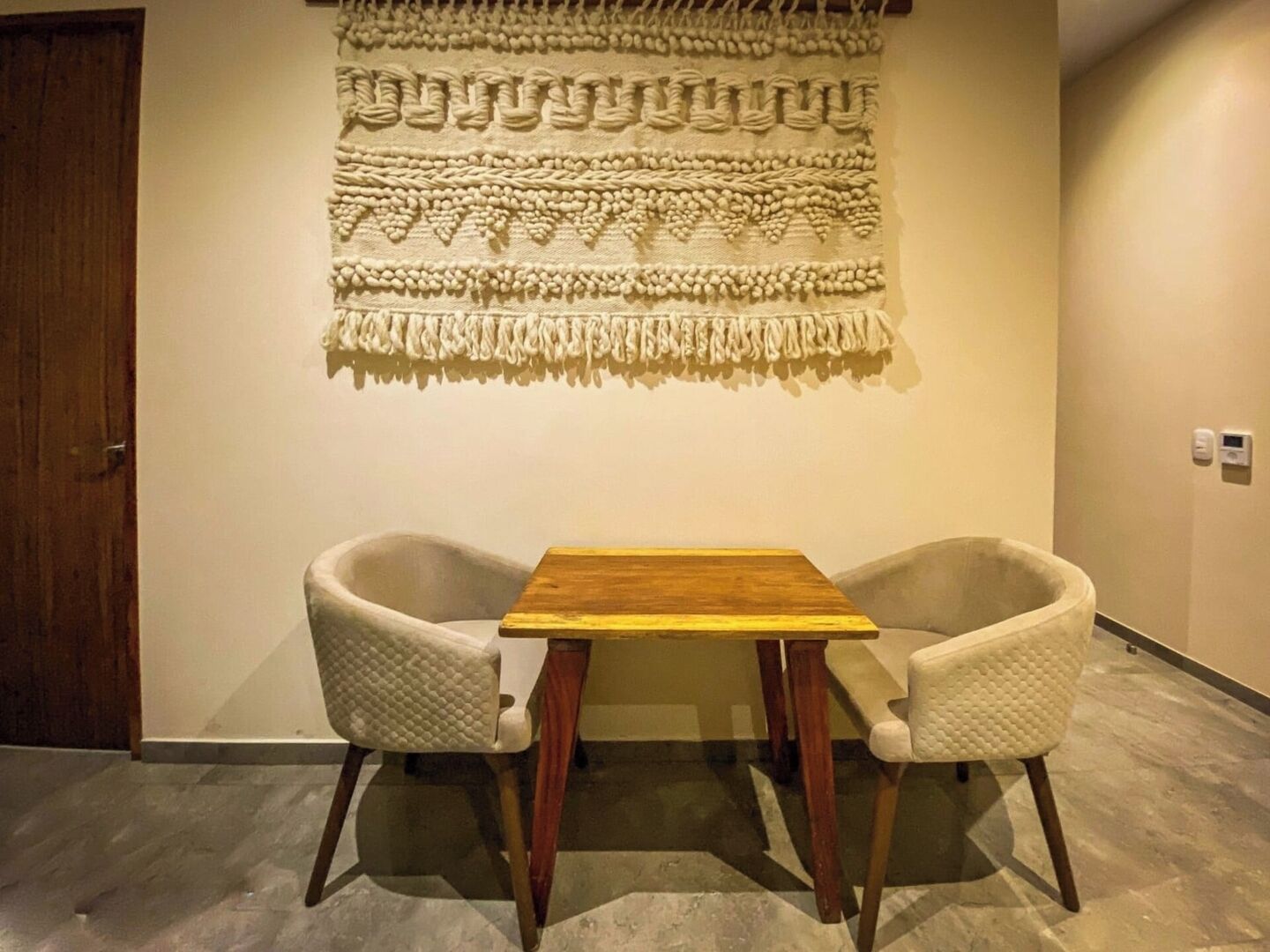 Una habitació amb una taula de fusta i dues cadires amb coixins, una obra de macramé penjada a la paret, i una porta de fusta.