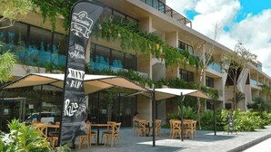 Edificio moderno con mesas al aire libre y una bandera que indica un bar.