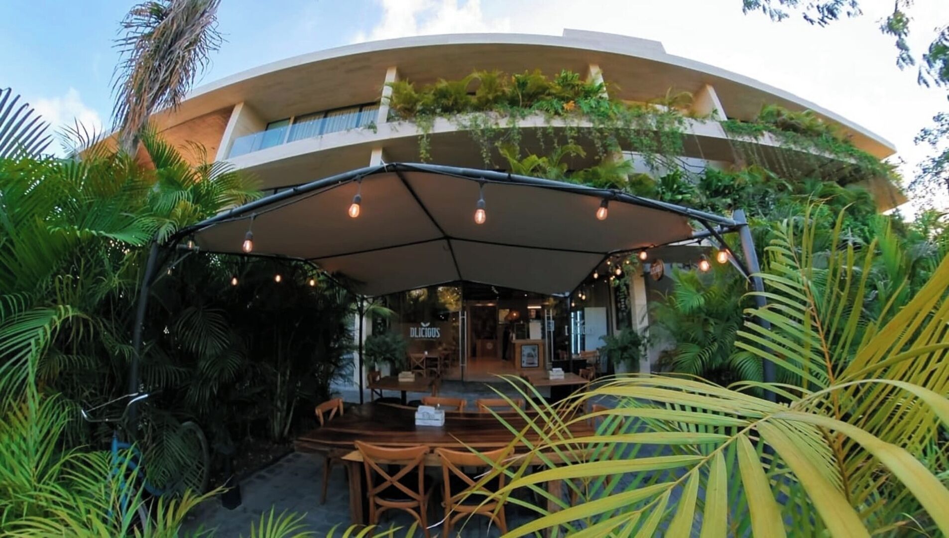 Un restaurante con terraza al aire libre cubierta por un toldo, rodeado de exuberante vegetación y un edificio moderno al fondo.