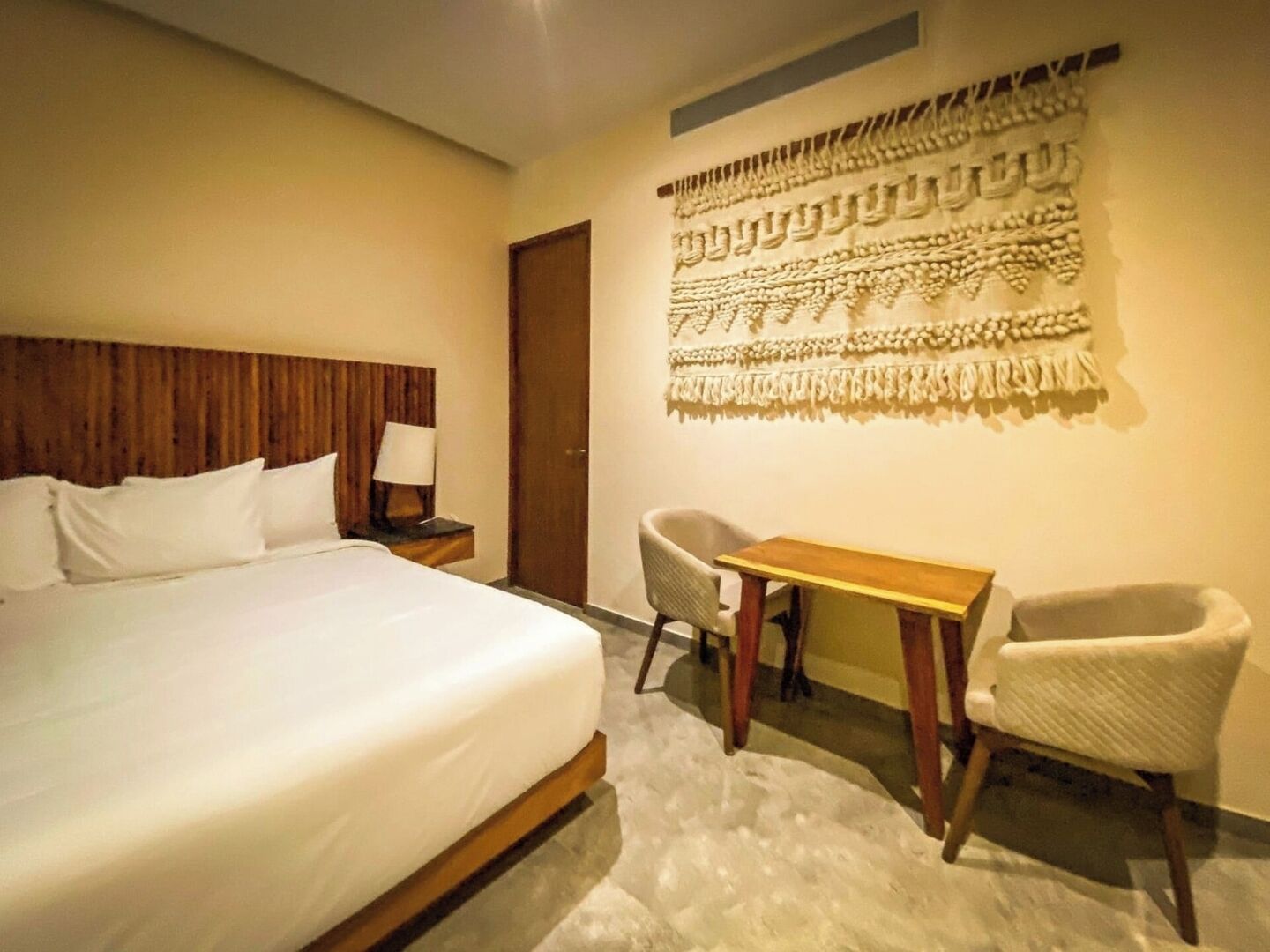A hotel room with a large bed, a small table, two chairs, a nightstand with a lamp, and a decorative wall hanging.