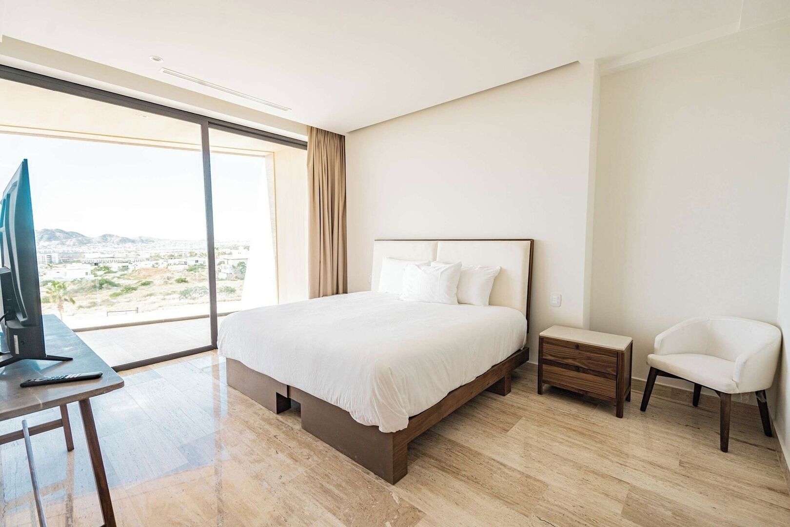 A modern hotel room with a large bed, a desk with a TV, a chair, and a view of the city.
