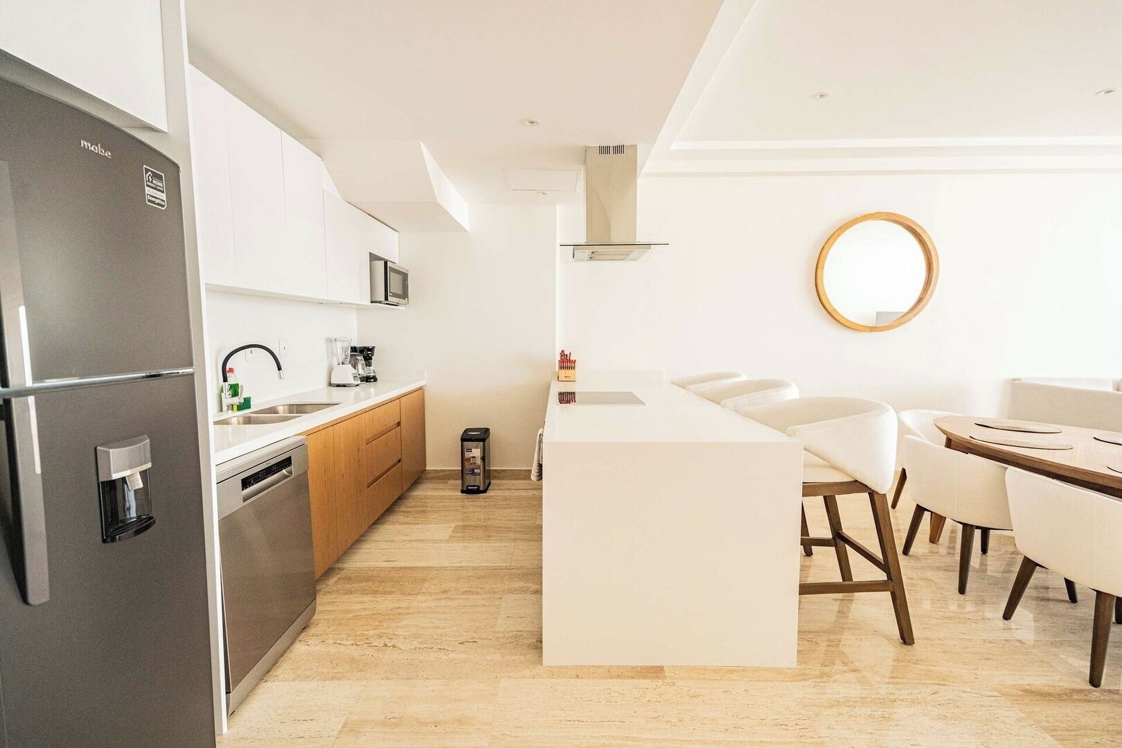 A modern kitchen with a built-in refrigerator, microwave, and coffee maker. There is a small trash bin and a round mirror on the wall.