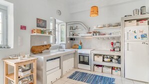 Una cocina moderna con armarios blancos, horno empotrado, lavamanos y estufa. Se observan estantes con vajilla y un lámpara colgante.