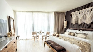 Quarto de hotel moderno com uma cama grande, televisão, área de refeições com cadeiras e uma janela ampla com cortinas.