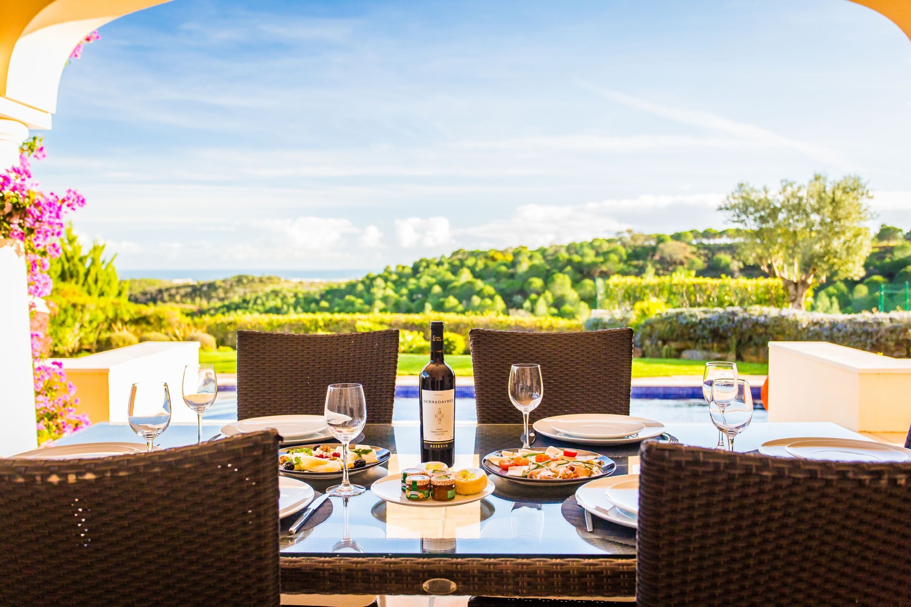 Mesa de comedor preparada para cuatro, con una botella de vino, copas y platos de comida, con vistas a un paisaje exuberante.