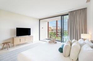 A modern hotel room with a large bed, a flat-screen TV on a wooden cabinet, a balcony with a view of buildings and greenery, and a lamp on the bedside table. - Top Floor Studio with Stunning Views and Balcony (Tulum)