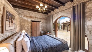 Un dormitorio con pared de piedra, techo de madera, cama con colcha estampada, candelabro y vista a través de un arco.
