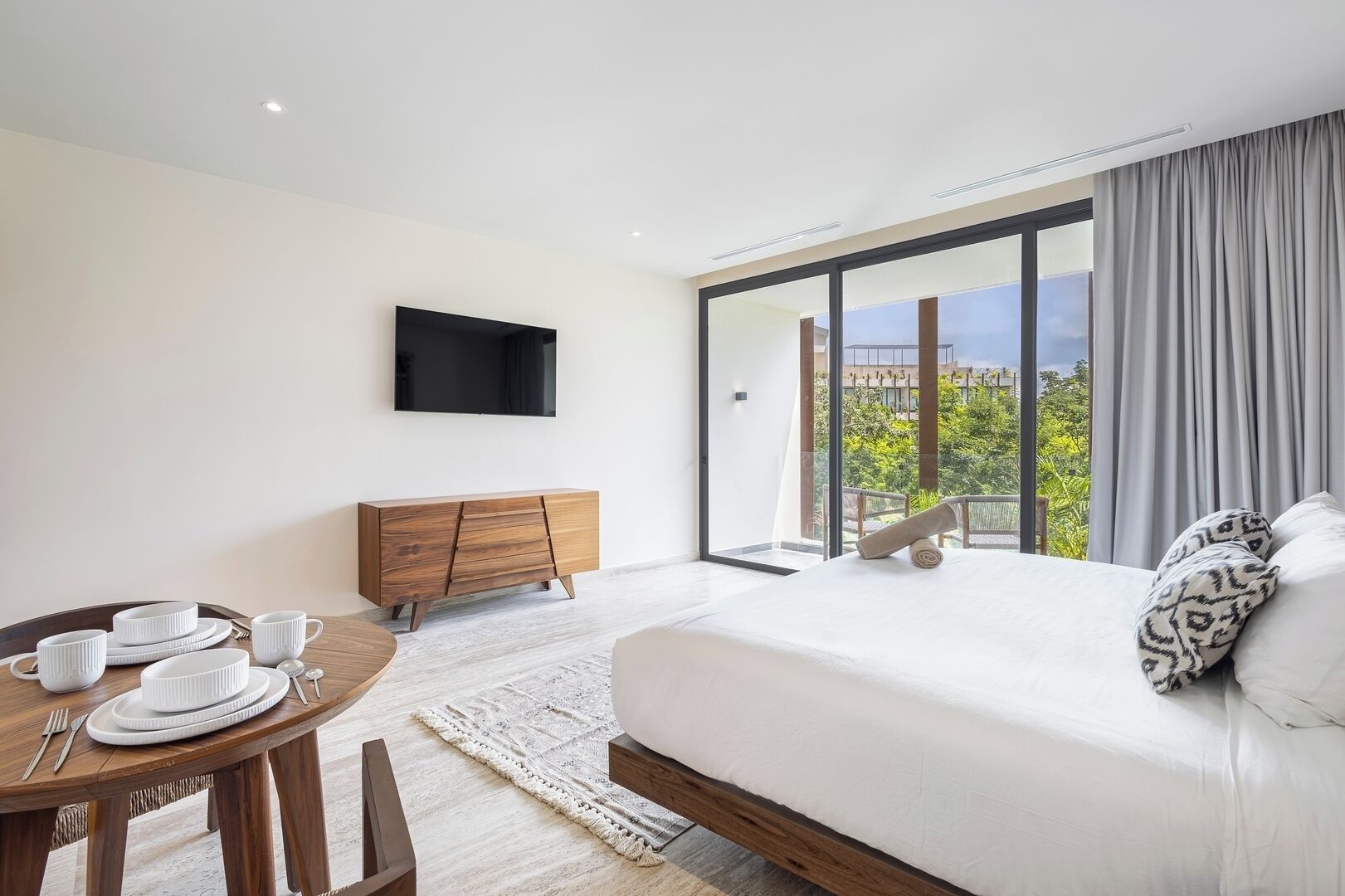 Quarto de hotel moderno com uma cama grande, área de refeições, cômoda de madeira e televisão na parede.