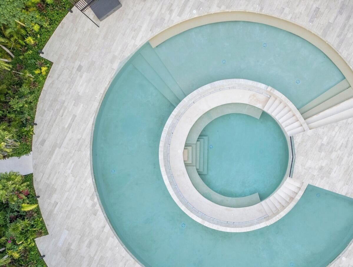 Une piscine circulaire dotée d’un escalier central et d’une série de marches périphériques, entourée d’une zone pavée agrémentée de verdure.
