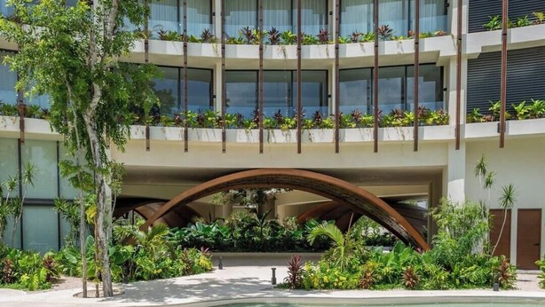 Een modern hotel met een zwembad, meerdere balkons met planten en een overdekte doorgang.