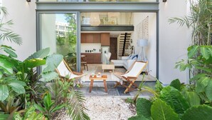 Uma casa moderna com porta de vidro que dá para uma área de jantar com duas cadeiras e uma mesa, cercada por vegetação exuberante.