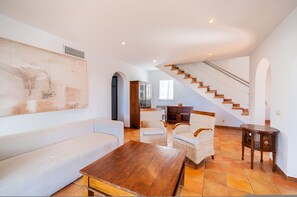 A spacious living area with a sofa, wicker chairs, a wooden coffee table, a staircase, and a large abstract painting on the wall. - Villa Finesse tipo 2 (Son Bou)