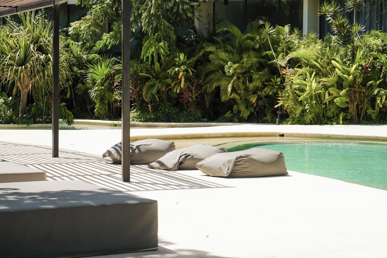 A poolside area with a lounge chair and cushions, surrounded by lush tropical plants.