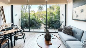A modern living room with a dining area, large windows, and a view of tropical plants. - Astounding Loft with Gym, Pool and Beach Shuttle (Tulum)