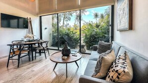 A modern living room with a dining area, large windows, and a view of tropical plants. - Astounding Loft with Gym, Pool and Beach Shuttle (Tulum)