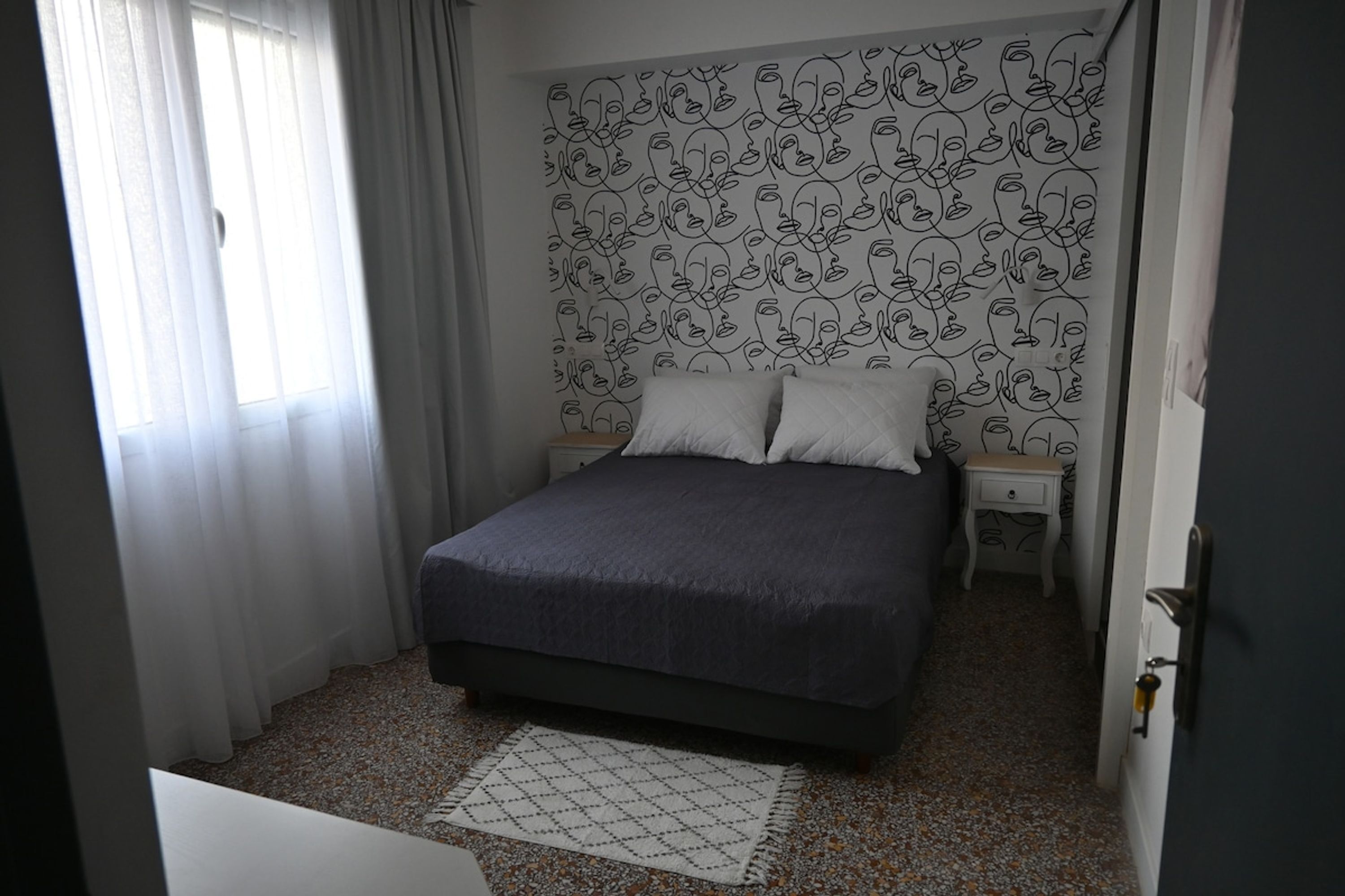 A bedroom with a bed, two bedside tables, a patterned wall, a window with curtains, and a door.