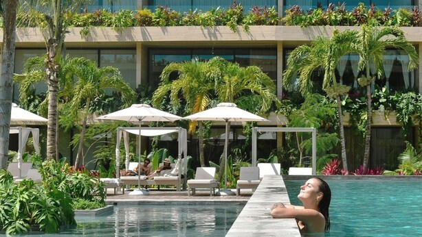 A pool area with lounge chairs and umbrellas, surrounded by lush greenery and a modern building facade. - Cozy Std, Steps to Infinity Pool, Shuttle to Beach (Tulum)