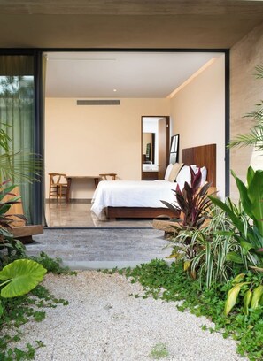 A modern bedroom with a large bed, a sitting area with a chair and table, and a view through a glass door. - Cozy Std, Steps to Infinity Pool, Shuttle to Beach (Tulum)