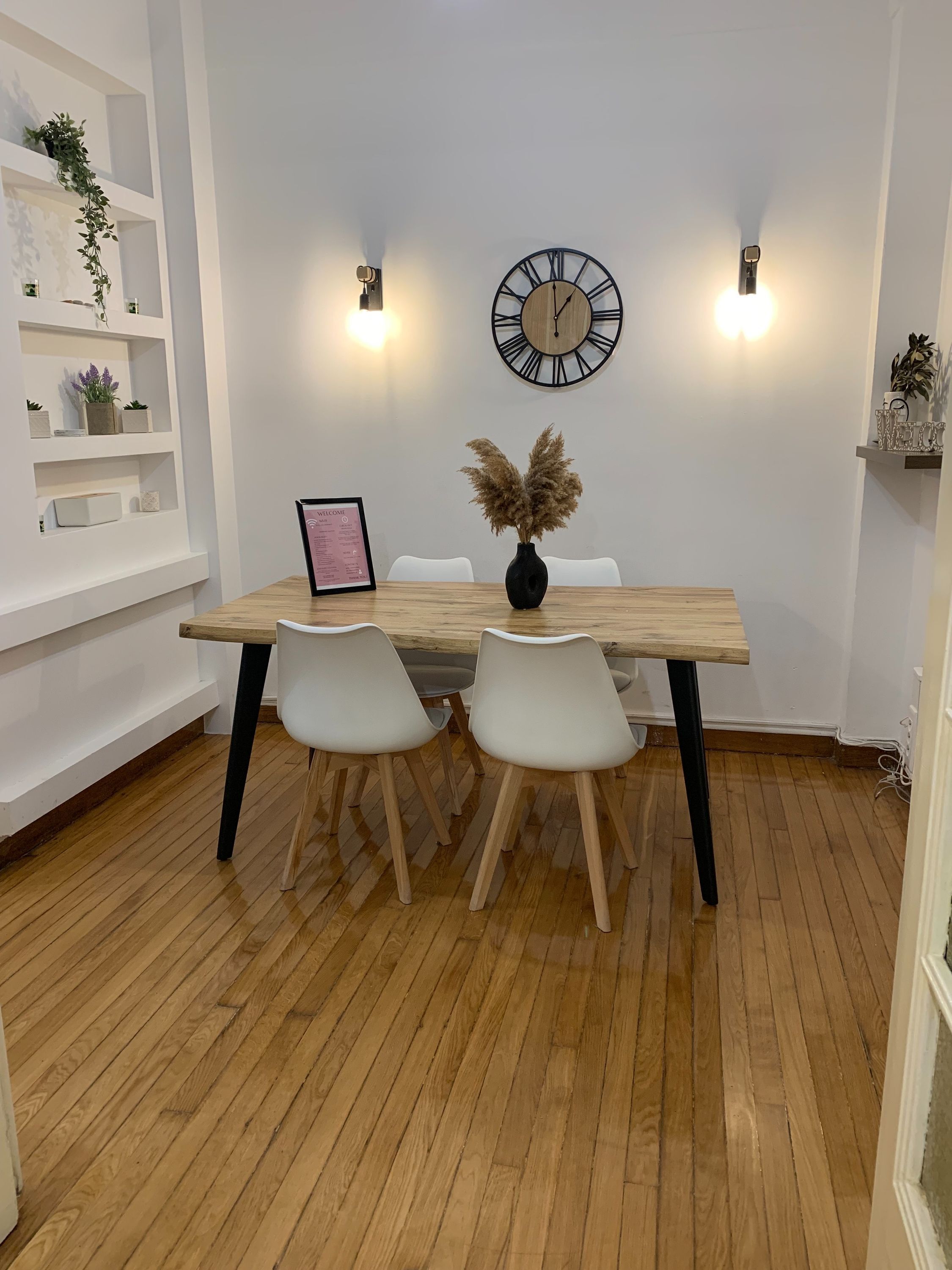 A dining area with a wooden table, white chairs, a clock, and wall-mounted lights.