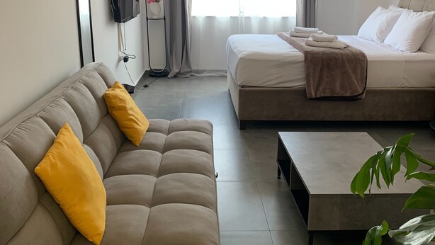 A modern hotel room with a sofa, a bed, a TV, and a large window with curtains. - Urban Hermes Apartment (Athina)