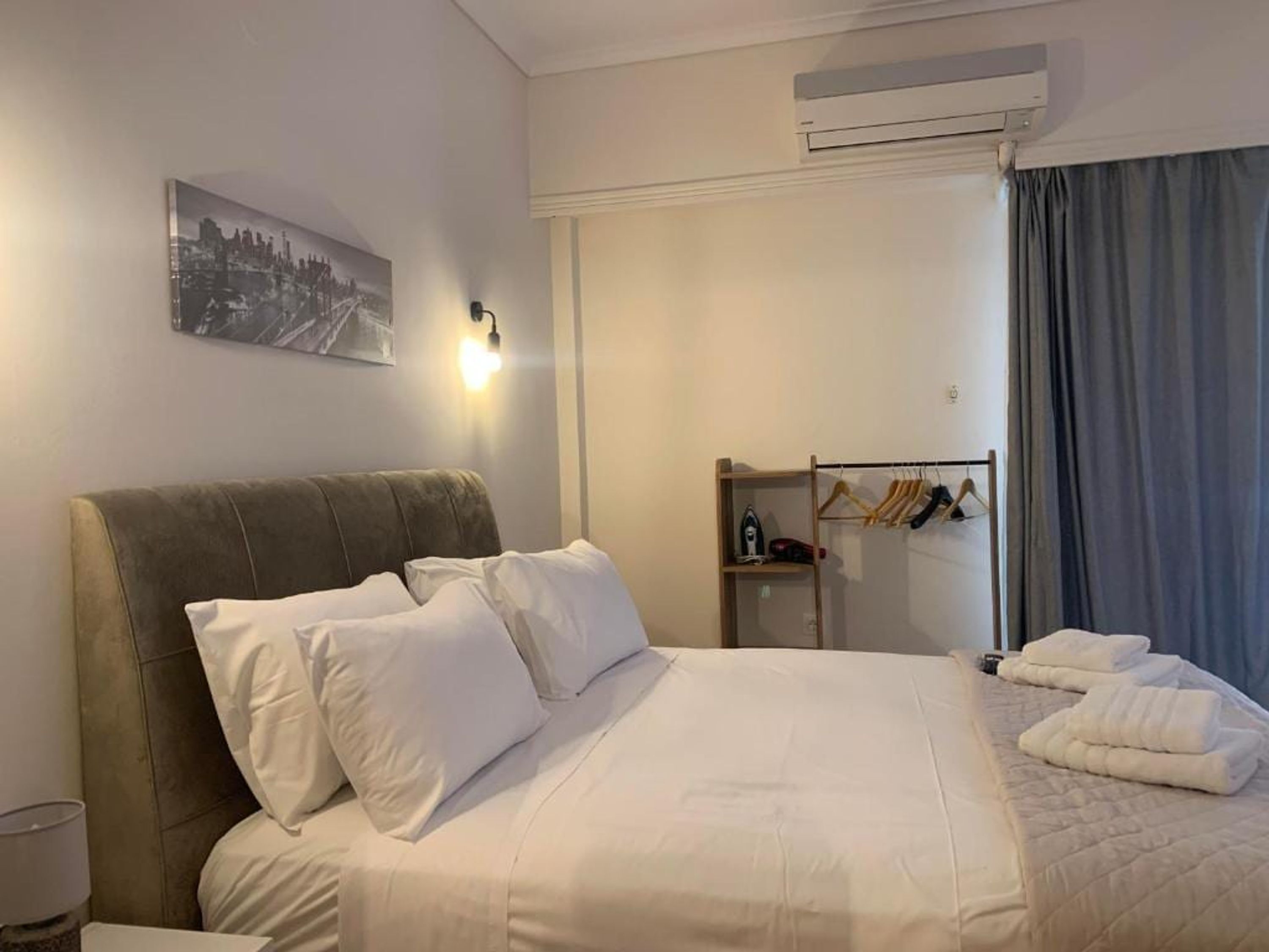 A hotel room with a bed, pillows, a nightstand, a mirror, a coat rack, and an air conditioning unit.