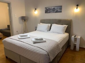 En snyggt bäddad säng med vita lakan och två hopvikta handdukar, en grå klädd sänggavel och en tavla med en stadsbild på väggen.