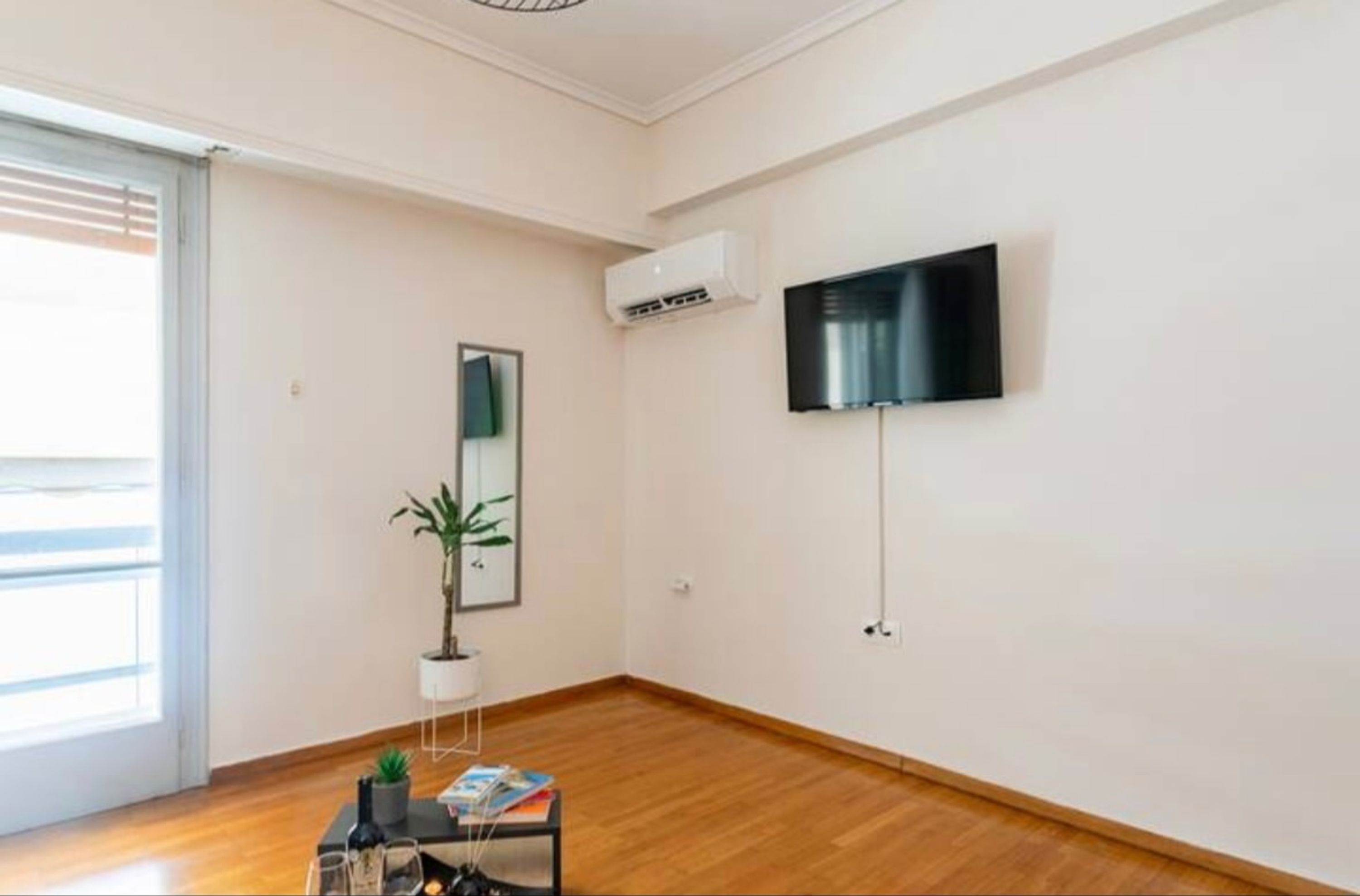 A minimalist living room with a wooden floor, a mounted flat-screen TV, a potted plant, and a small table with a magazine.