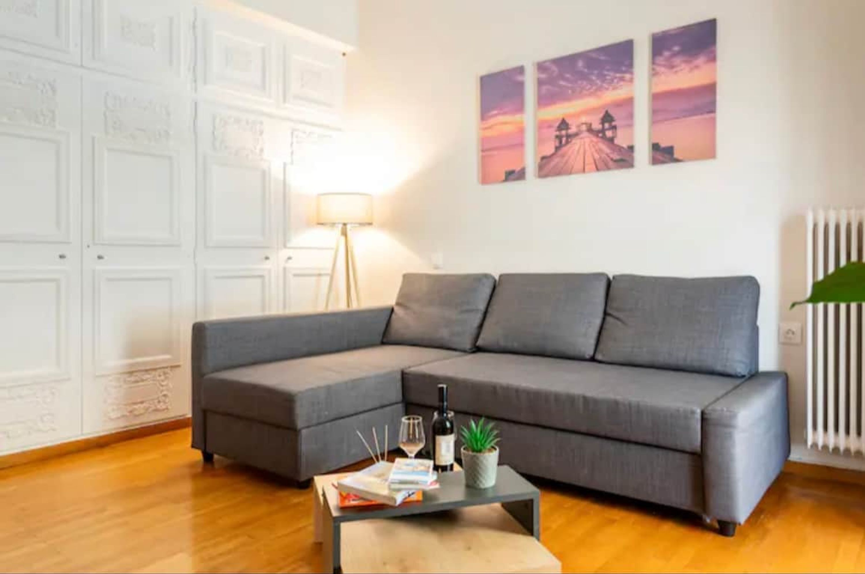 Ein modernes Wohnzimmer mit einem grauen Ecksofa, einem Couchtisch mit Büchern und einer Flasche, und einer Wand mit drei Sonnenuntergangsbildern.