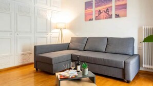 Un salon moderne avec un canapé d’angle gris, une table basse sur laquelle sont posés des livres et une bouteille, et un mur orné de trois tableaux représentant des couchers de soleil.