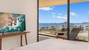 Slaapkamer met een groot raam met uitzicht op de kust, een schilderij aan de muur en een bed met een wit laken.