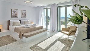 A modern bedroom with a large bed, a view of the outdoors, and a seating area with a chair and a table. - Aegean Hestia Luxury Villa of Paros (Paros)