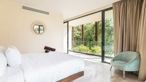 A bedroom with a large bed, a round mirror, a chair, and a sliding glass door leading to a balcony with greenery. - Fast Wi-Fi, private pool, A/C and jungle view! (Tulum)