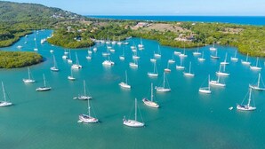 An aerial view of a marina with numerous sailboats docked in turquoise waters, surrounded by lush greenery and a distant coastline.