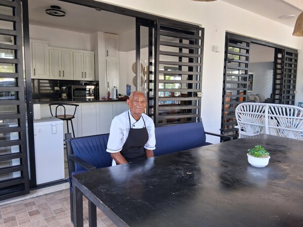A person seated at a table with a plant on it, in a room with a kitchen area and a white chair.