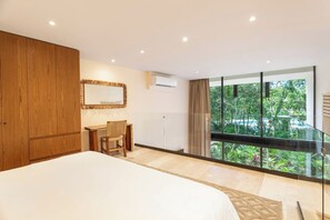 Ein Schlafzimmer mit einem großen Bett, einem Holzschrank, einem Schreibtisch mit Stuhl, einem Fenster mit Blick auf Grünflächen und einer Klimaanlage.