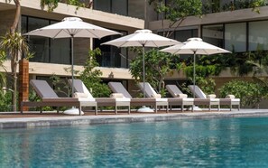 A poolside area with lounge chairs and umbrellas, surrounded by greenery and a modern building facade. - Perfect for family trips! 2 bedroom 3 beds AP (Tulum)