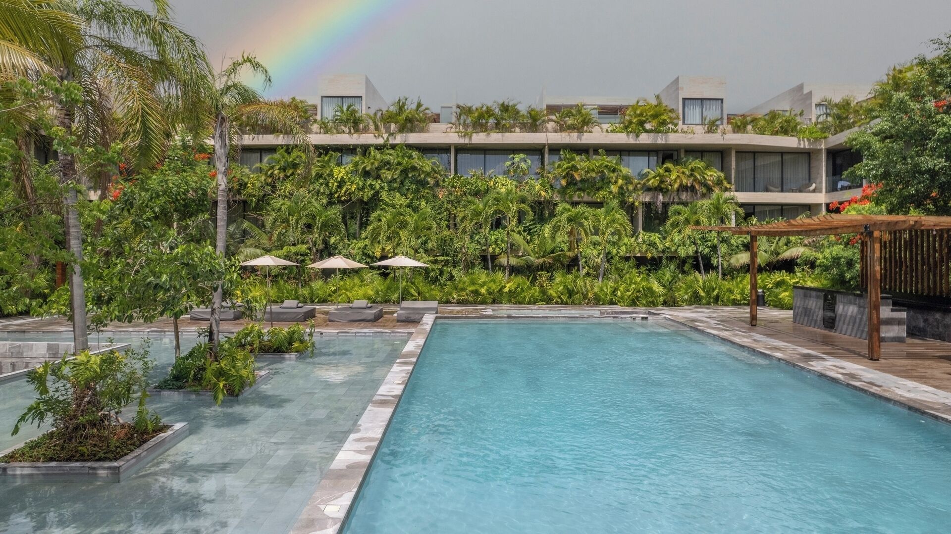 A swimming pool with lounge chairs and umbrellas, surrounded by lush greenery and modern buildings.