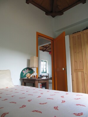Een slaapkamer met een bed, een houten garderobe, een houten tafel met een ventilator en lamp, en een spiegel.