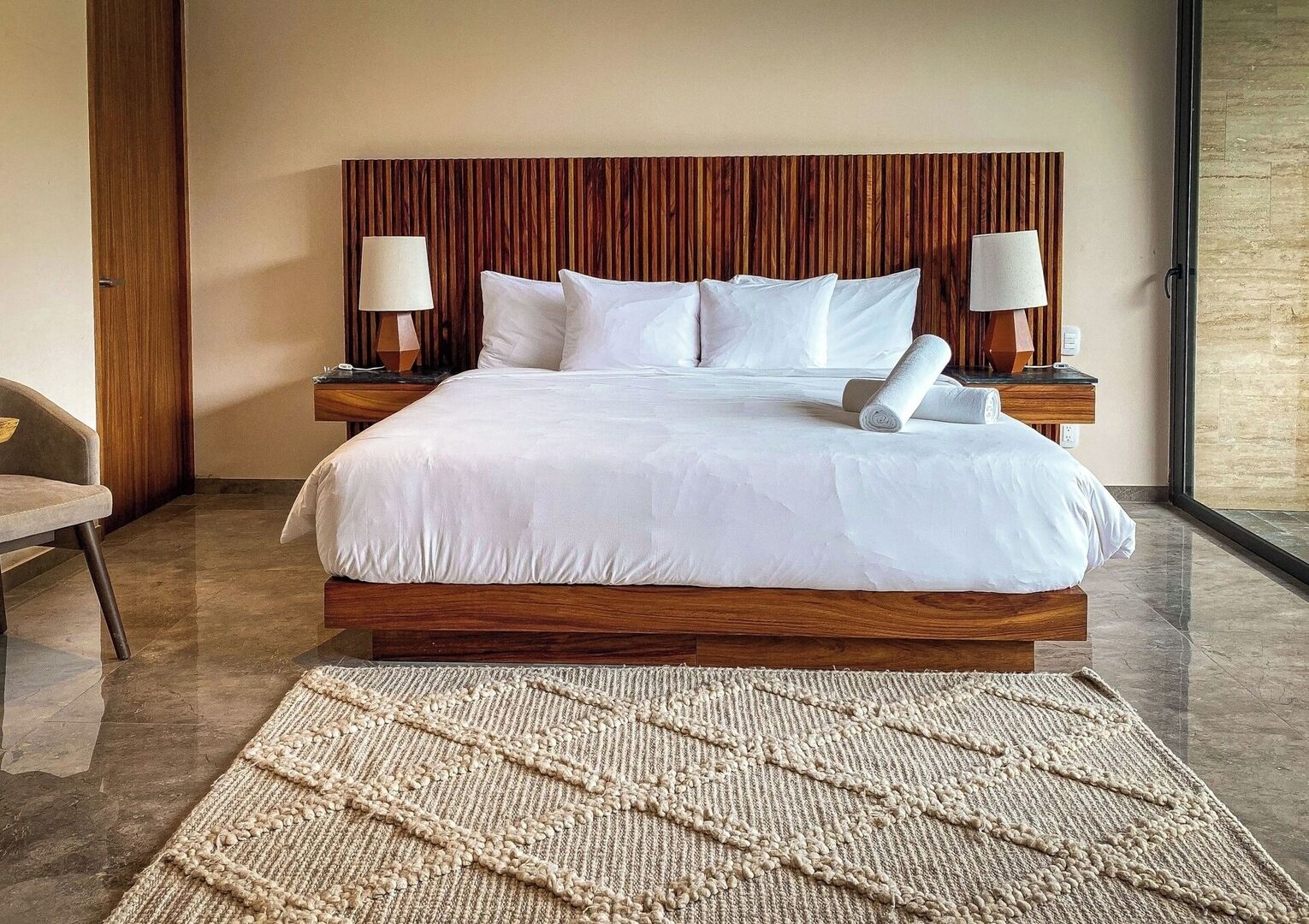 Ein modernes Hotelzimmer mit einem großen Bett, einem Holzkopfteil, zwei Nachttischen mit Lampen, einem Sessel und einem großen Teppich.