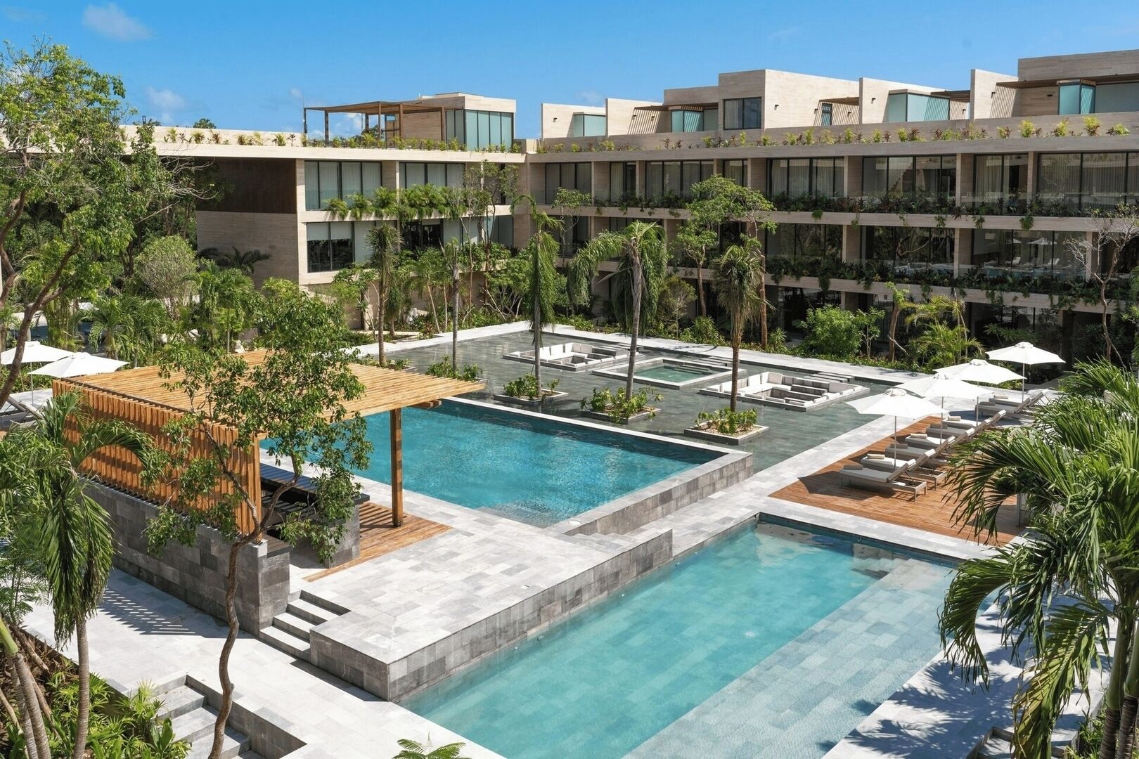 A modern hotel with multiple swimming pools, outdoor seating areas, and lush greenery.