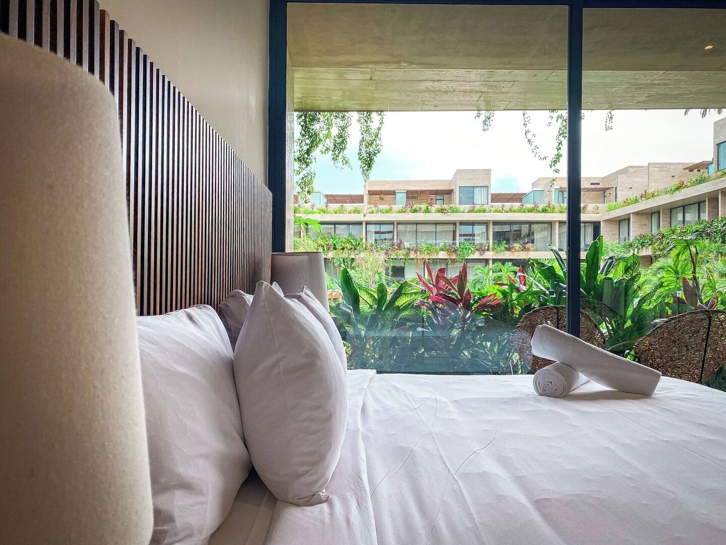 Ein modernes Hotelzimmer mit einem großen Bett, weißen Leinen und Blick auf einen üppigen Innenhof mit Pflanzen und Gebäuden.