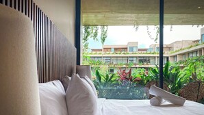 Une chambre d’hôtel moderne avec un grand lit, du linge de lit blanc et une vue sur une cour verdoyante agrémentée de plantes et de bâtiments.
