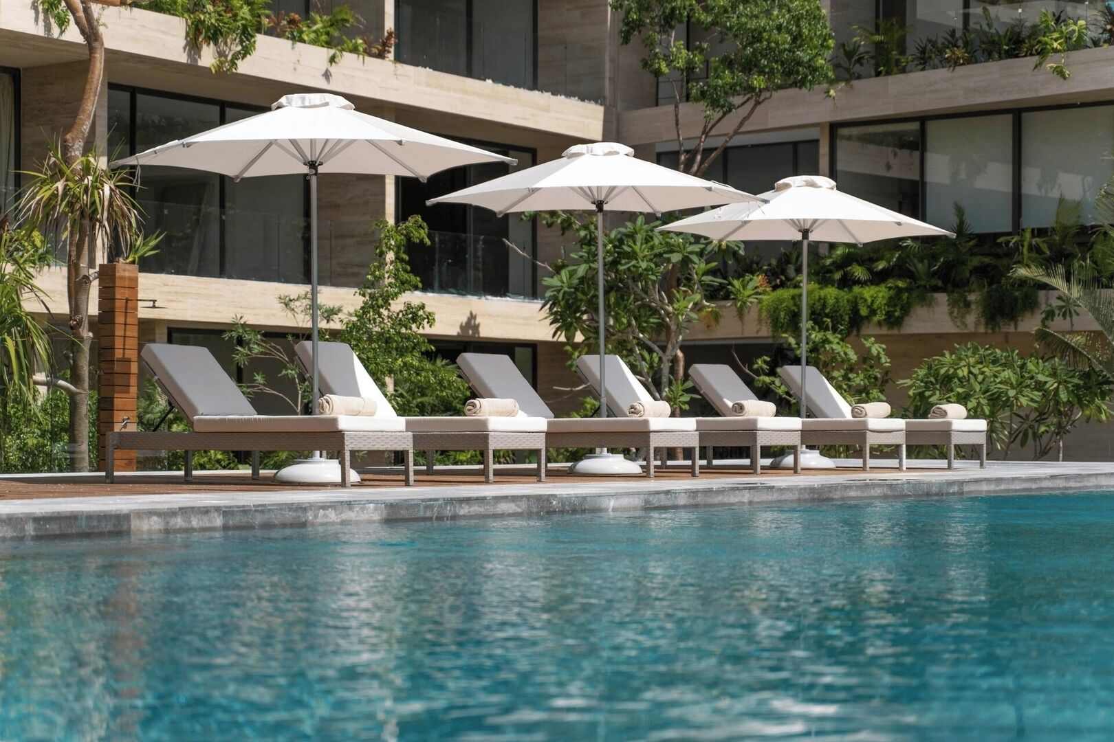 Un espace aménagé au bord de la piscine, avec des chaises longues et des parasols, entouré d’un bâtiment doté de grandes fenêtres et agrémenté de verdure.