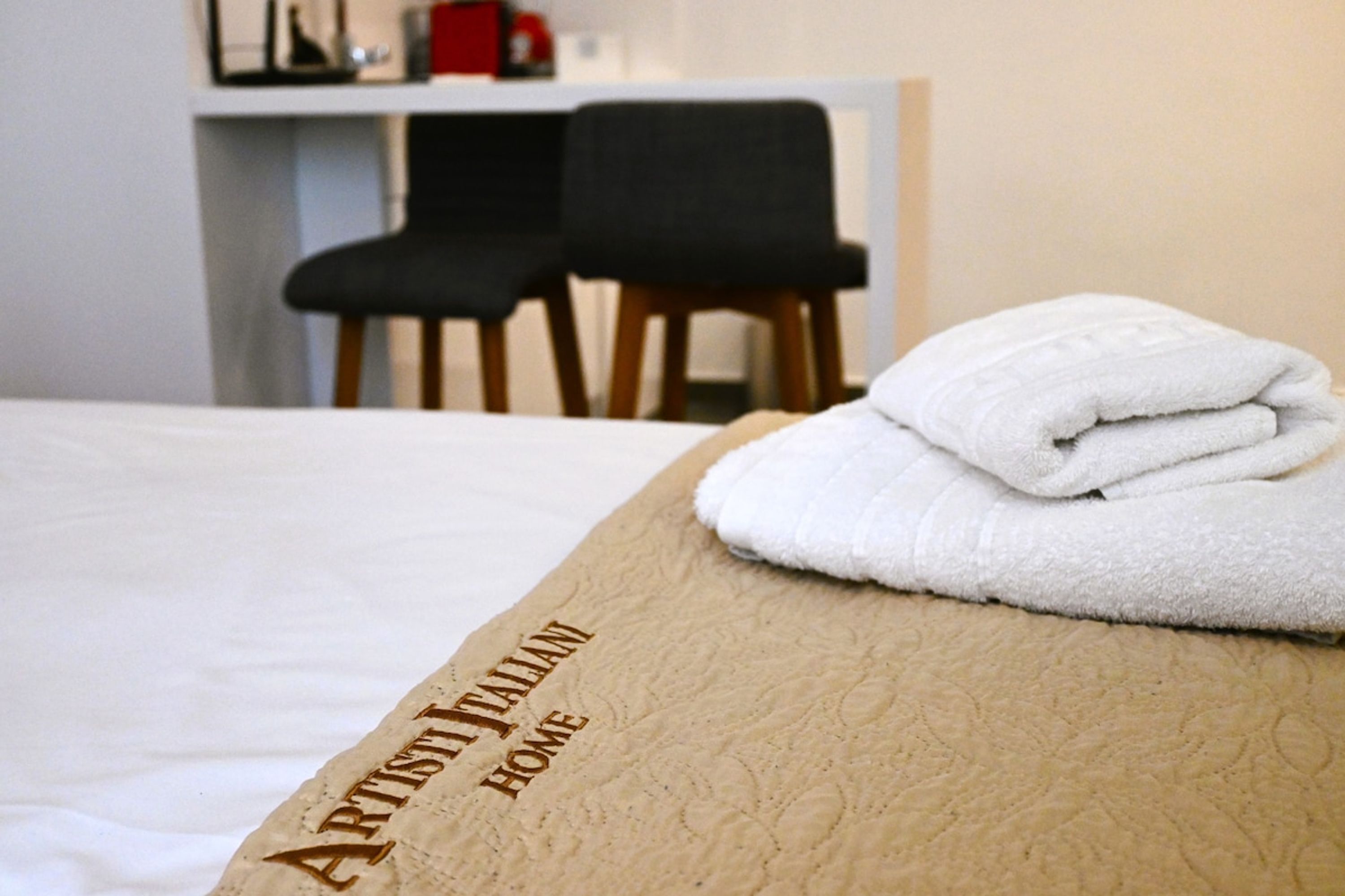 Ein ordentlich bezogenes Bett mit einem gefalteten weißen Handtuch am Kopfteil und einem braunen Kissen mit dem Aufdruck "ARTISTY HOME".