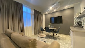 Ett modernt hotellrum med en stor soffa, en matplats med bord och stolar, en platt-TV som är monterad på väggen och ett litet kök.